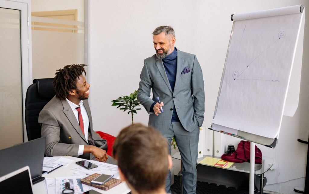 A business leader smiles while presenting beside a flip chart to colleagues gathered around a conference table.