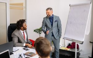 A business leader smiles while presenting beside a flip chart to colleagues gathered around a conference table.