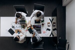Overhead view of four colleagues joining hands across a conference table covered with laptops, charts and work materials.